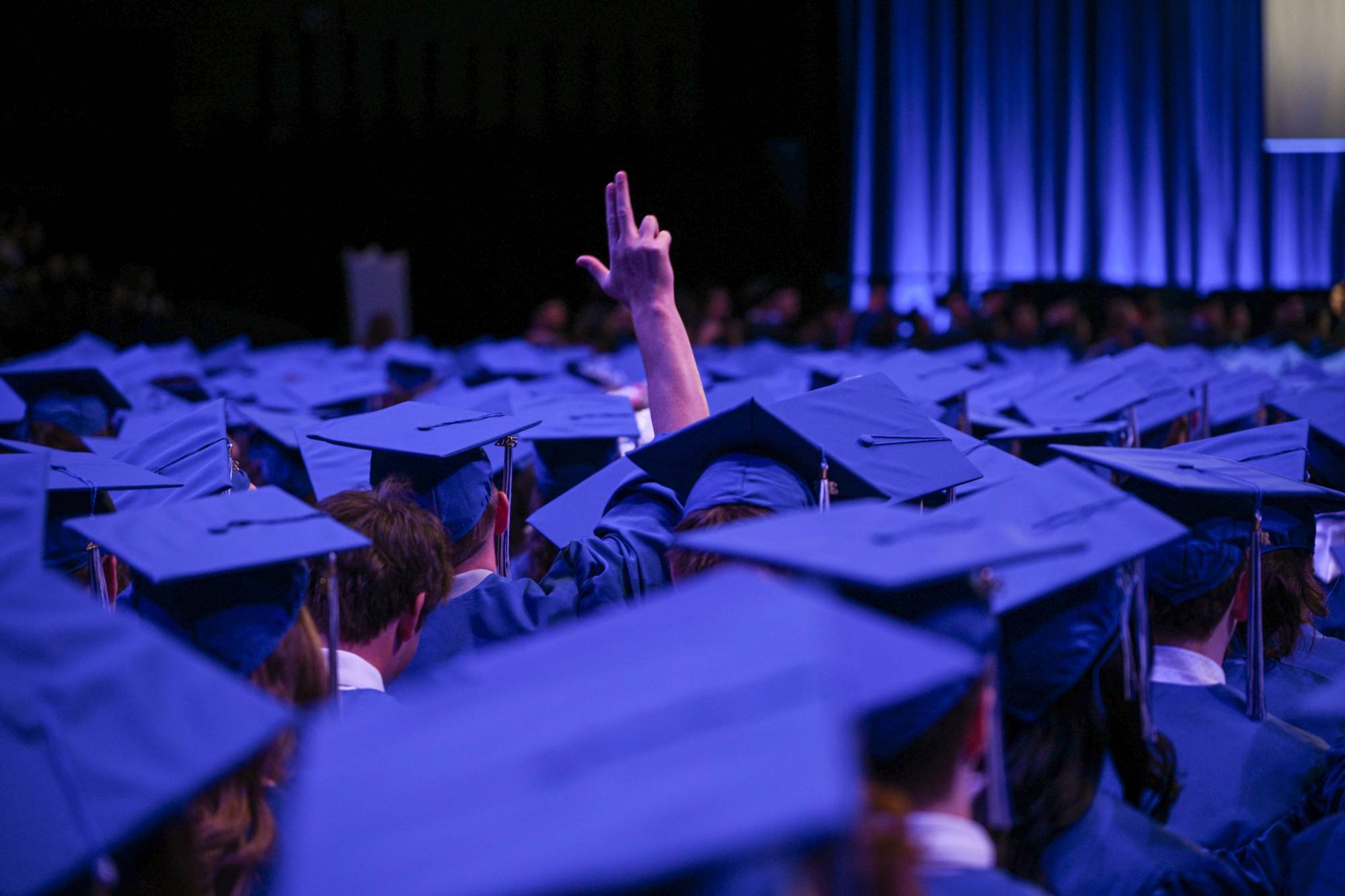 Aerial view of several GVSU graduates wearing blue caps and one of them raising their hand giving the anchor up sign